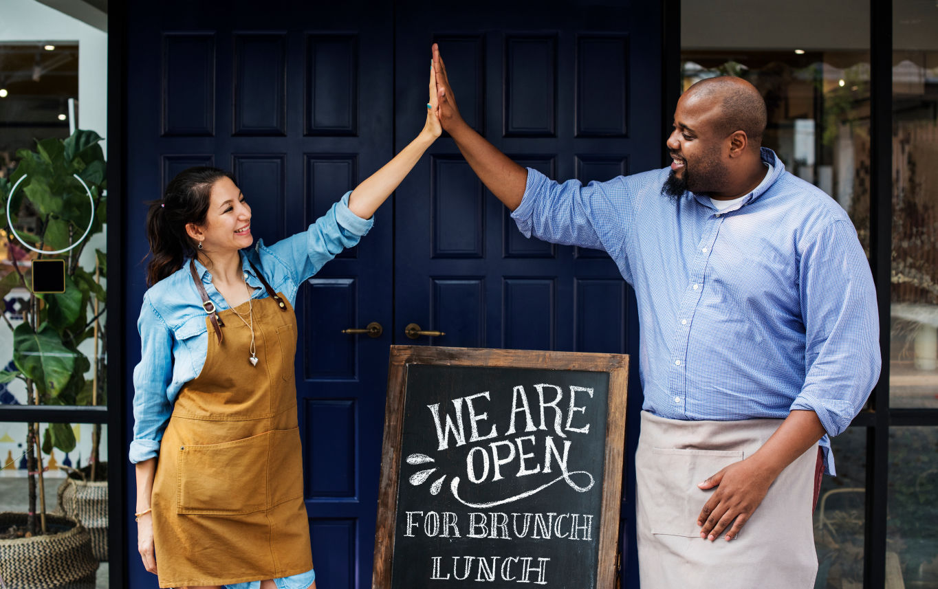 man and woman high five in front of a store front with open sign